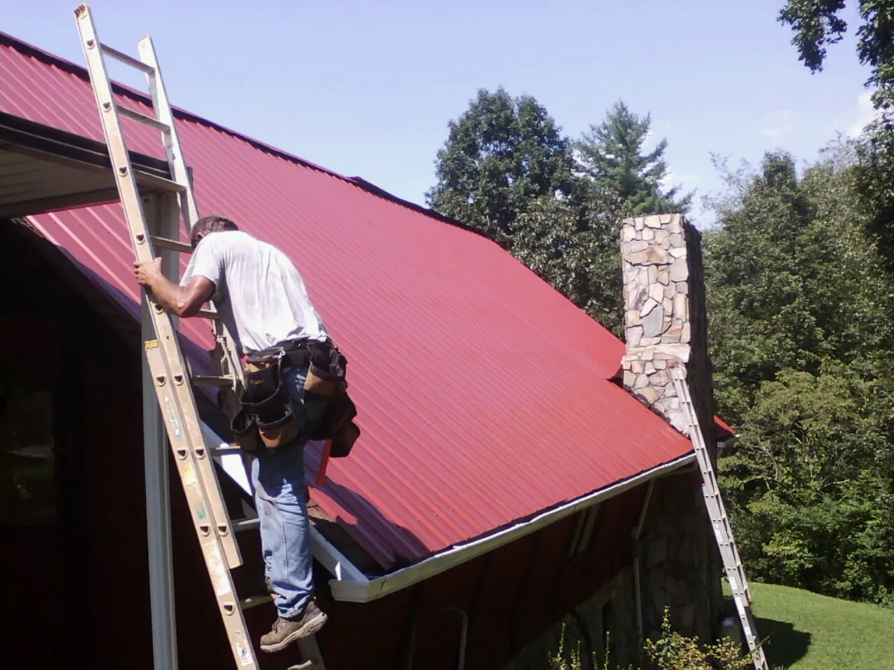 Roofer on a ladder beside a red metal roof and stone chimney — Soffit & Fascia Repair by Roofing Brothers in Bally